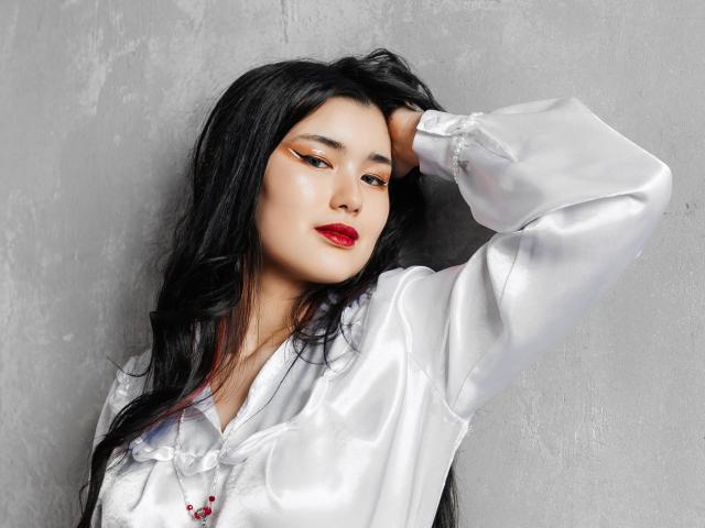 AnneTLuis with long black hair, white satin shirt, red lipstick, and winged eyeliner, arm raised, hand in hair, looking slightly to the side, sexy and confident.