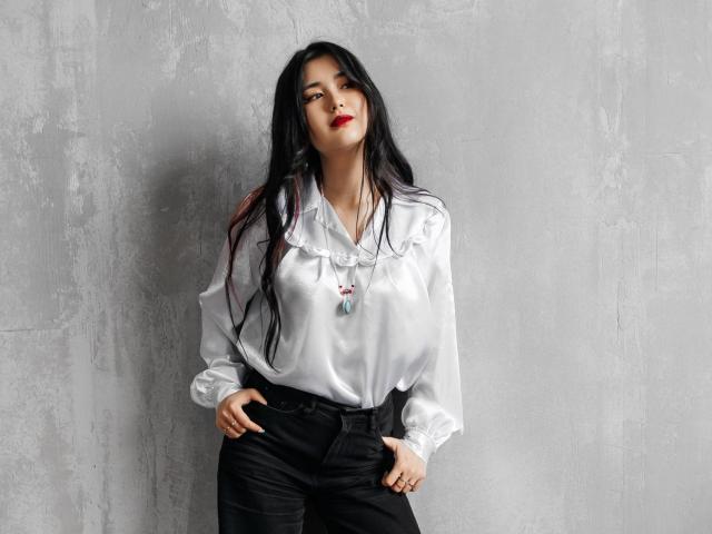 AnneTLuis with long black hair, wearing a white satin blouse, black pants, and a colorful necklace, standing against a grey textured wall, hands in pockets, red lipstick.