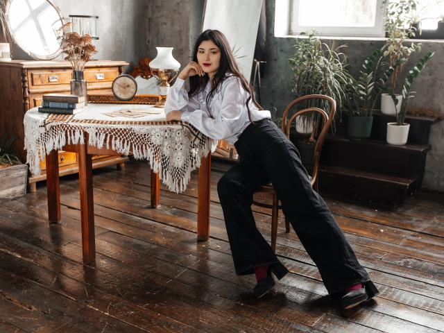 AnneTLuis leans on a wooden table, wearing a white shirt, black pants, and red socks, with her ass slightly raised, in a rustic room with a fringed tablecloth.
