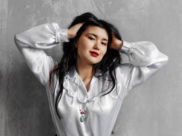 AnneTLuis with long black hair, wearing a shiny white blouse, hands in her hair, red lipstick, and a necklace with a blue pendant, smiling subtly.