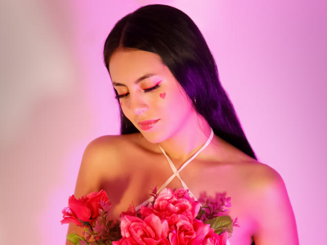 EimmyPark with long black hair, pink heart makeup, eyes closed, holding red roses, criss-cross white necklace, pink and purple lighting, soft smile, upper body, bare shoulders.