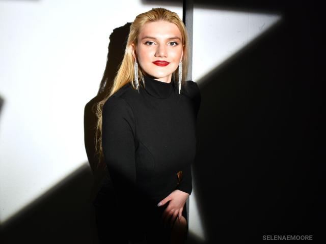 SelenaeMoore with long blonde hair, wearing a black turtleneck, standing in shadow, smiling with red lipstick, hands clasped, light highlighting her face and hair.