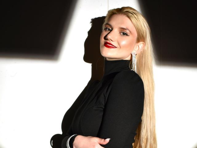 SelenaeMoore, avec de longs cheveux blonds, un pull noir à col roulé, du rouge à lèvres, et les bras croisés, souriante, avec une ombre sur le mur blanc, des boucles d'oreilles, et la lumière du soleil mettant en valeur son visage. Ses seins sont légèrement visibles sous le pull à col roulé..