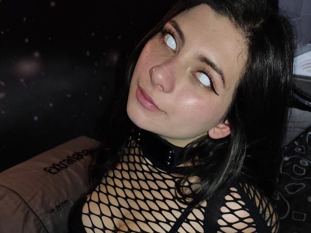 JeneKurz with pale skin, dark hair, and glowing blue eyes, wearing a black fishnet top and a spiked choker, smiling slightly. Boobs visible through the fishnet.