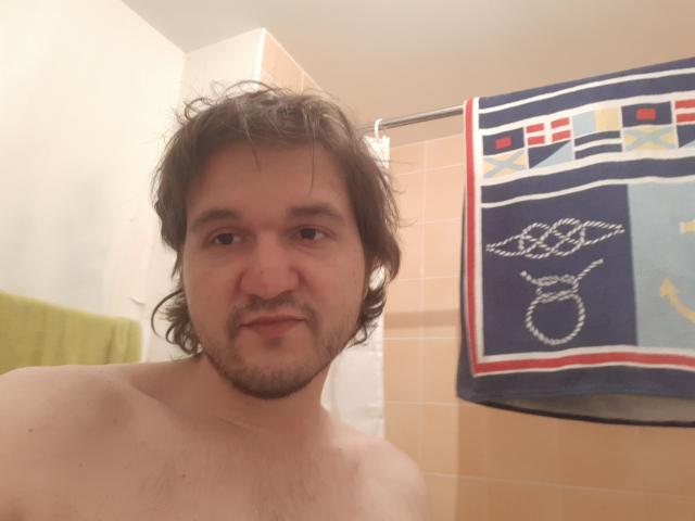 NectarineFad is a shirtless dude with wavy brown hair, a beard, and a nose ring, standing in a bathroom with a towel hanging on a shower curtain.