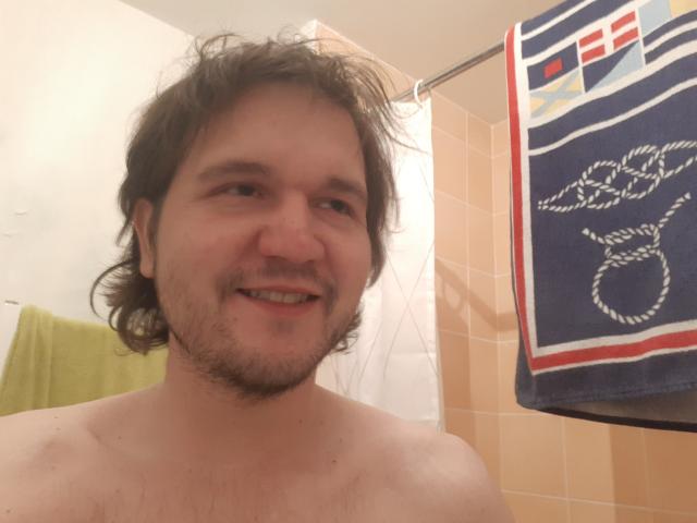 NectarineFad, shirtless, with wavy brown hair, beard, and a slight smile, standing in a bathroom, holding a blue towel with white and red patterns.