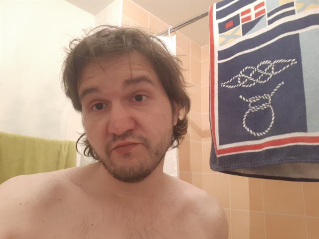 NectarineFad has a slightly chubby, shirtless face with messy brown hair, a beard, and a bored expression in a bathroom. A green towel and a blue towel hang nearby.