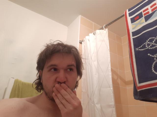NectarineFad has shoulder-length brown hair, a beard, and is shirtless, covering his nose with his hand in a bathroom. A blue towel with a red and white design hangs.