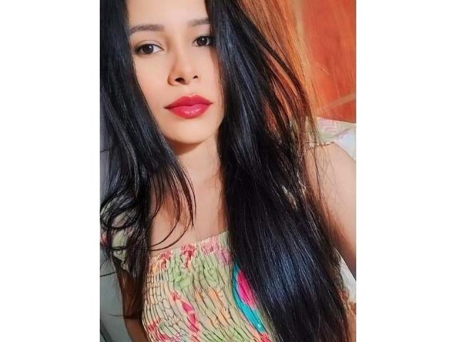 AnandaHot with long black hair, red lipstick, and a colorful, striped top, showing off her smooth, light brown skin and subtle makeup. Her small tits are partially visible.