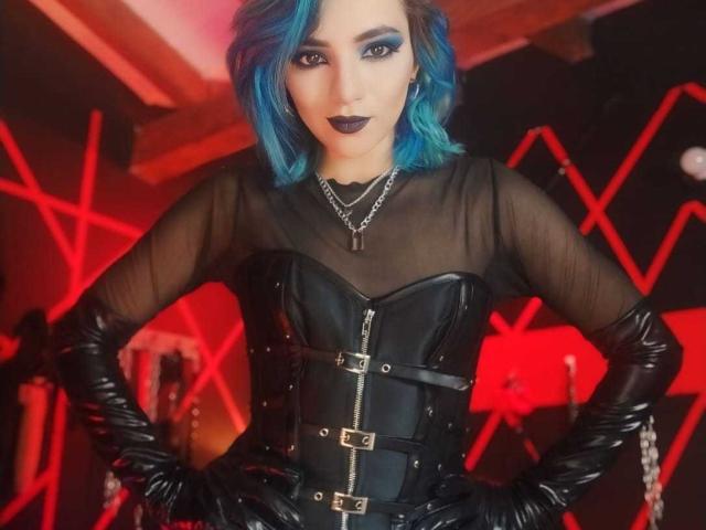 MisssDarkLady with blue hair, black sheer top, leather corset with buckles, and black gloves, hands on hips, dark lipstick, and chain necklace, standing confidently.