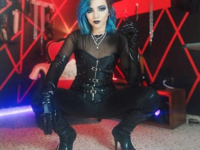 MisssDarkLady with blue hair, black sheer top, leather corset, gloves, and boots, squatting with legs spread, wearing black lipstick, and a cross necklace.