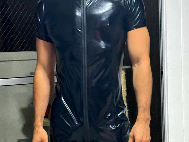 RebelDali69 is wearing a shiny black latex bodysuit, zipped up, with muscular arms and no visible genitals or boobs. The bodysuit is tight and reflective.