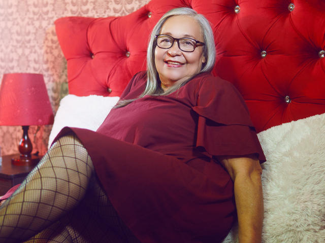 RosarioTriana with gray hair, glasses, red dress, and fishnet stockings, smiling on a red tufted bed, legs crossed, red lamp on the left. Her ass is partially visible.