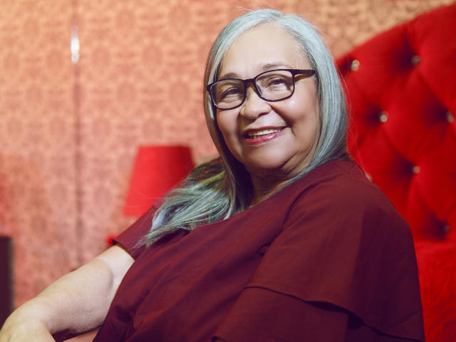 RosarioTriana with gray hair, glasses, and a red shirt, smiling with her tits slightly visible, sitting on a red tufted chair, red lamp in the background.