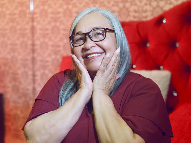 RosarioTriana with blue hair, glasses, smiling, hands up to cheeks, wearing a maroon shirt, red tufted headboard, and pinkish wall in the background.