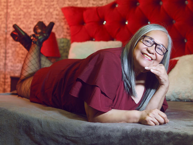 RosarioTriana with gray hair, glasses, red dress, lying on bed, smiling, hand under chin, fishnet stockings, red-tufted headboard, gray bed, black heels, sexy, relaxed.