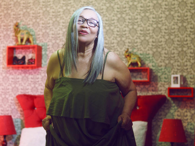 RosarioTriana, with gray-green hair and glasses, pulling down a green dress to reveal her large, bare, sagging tits. She's smiling, standing in a colorful, patterned room.