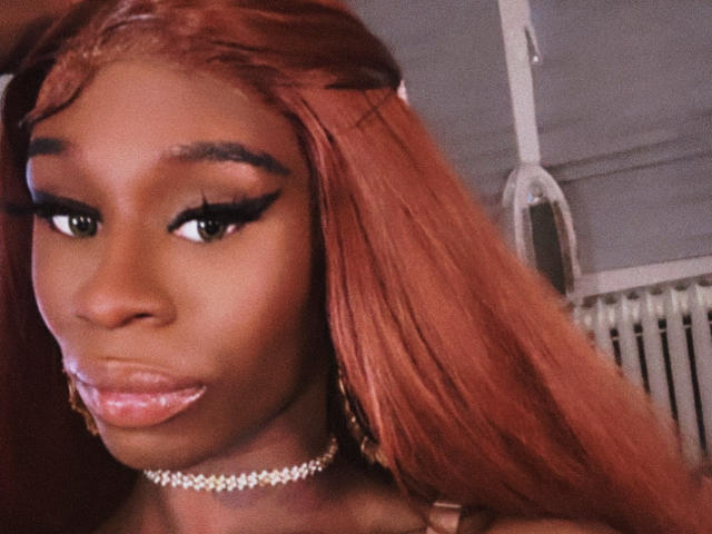 TsHottieX has dark brown skin, long red hair, thick black eyeliner, shiny pink lips, a diamond choker, and gold earrings, looking down with a serious expression.