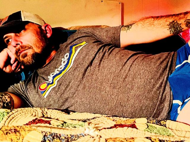 Waldrip, a bearded dude with tattoos, laying on a bed in a grey t-shirt and blue shorts, wearing a backwards cap, with a thoughtful expression, and a quilted blanket.