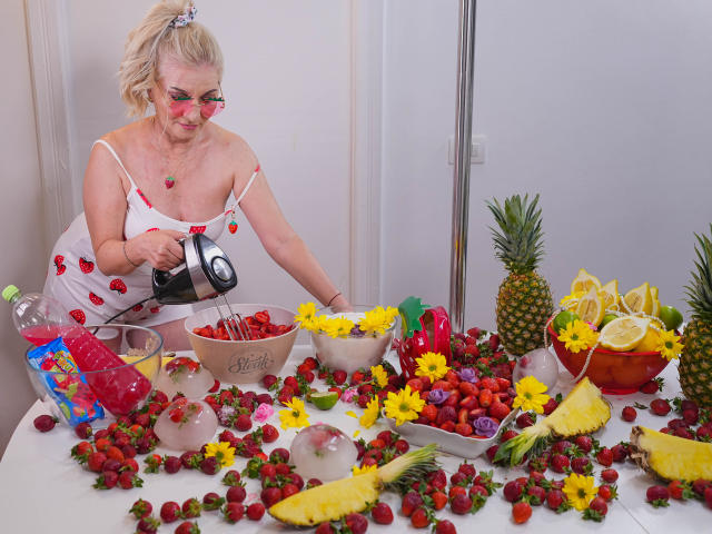 LadyJanyne with blonde hair, red face paint, and white strawberry dress, pours from a black teapot into a bowl of strawberries, surrounded by pineapple, lemon, and fruit.