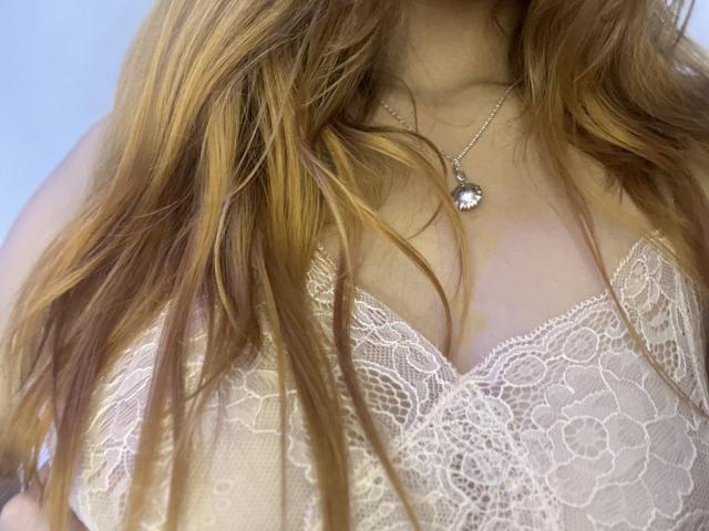 ShantalSummer with long, wavy blonde hair, wearing a white lace bra that shows her small, perky tits. She's got a silver necklace with a round pendant.