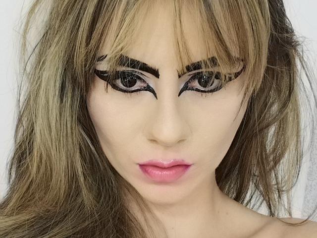 KariCarter's face with blonde, wavy hair, thick black eyeliner, and pink lipstick. Her eyebrows are bold, and her eyes are looking directly at the camera.