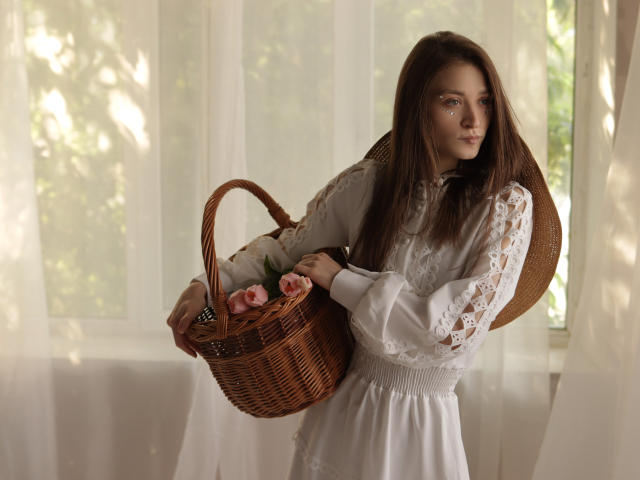 MiaBerry with long brown hair, wearing a white lace dress, holds a wicker basket of pink roses, standing near a sunlit window, looking slightly to the side.