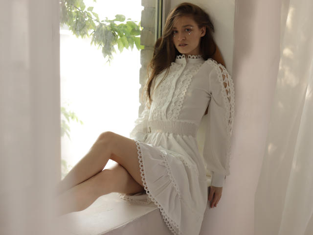 MiaBerry with long brown hair, wearing a white lace dress, sitting on a window ledge, legs crossed, looking at the camera with a soft expression, light filtering through.