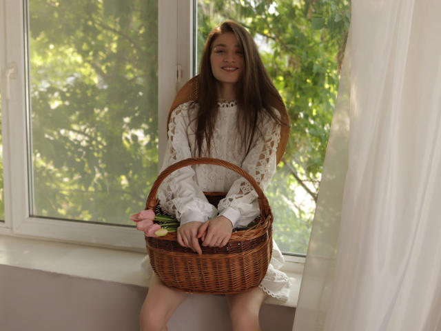 MiaBerry with long brown hair, wearing a white lace dress, sitting on a windowsill, holding a wicker basket with pink flowers, smiling, white curtain on the right.