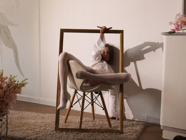 MiaBerry with pale skin and curly blonde hair, wearing a white knit robe and white heels, sitting in a modern chair, legs spread, holding a wooden frame.