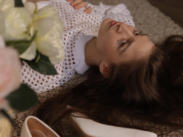MiaBerry with long brown hair, lying on a textured surface, wearing a white crochet top, eyes adorned with glitter, white peony flowers near her head, pink shoe in foreground.