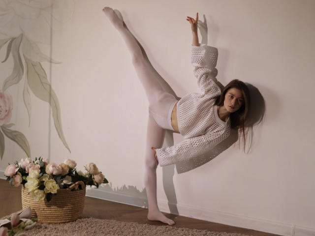 MiaBerry, with white tights and crocheted top, bends back, raising one leg high, showing her ass and small tits, against a white wall, with a basket of flowers.