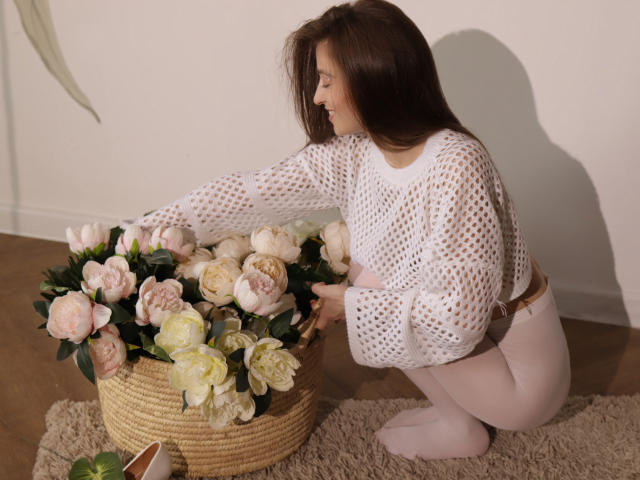 MiaBerry, squatting, wearing white mesh top and pink tights, touching a wicker basket filled with pink and white roses. Brown hair, soft light, wooden floor, beige rug.
