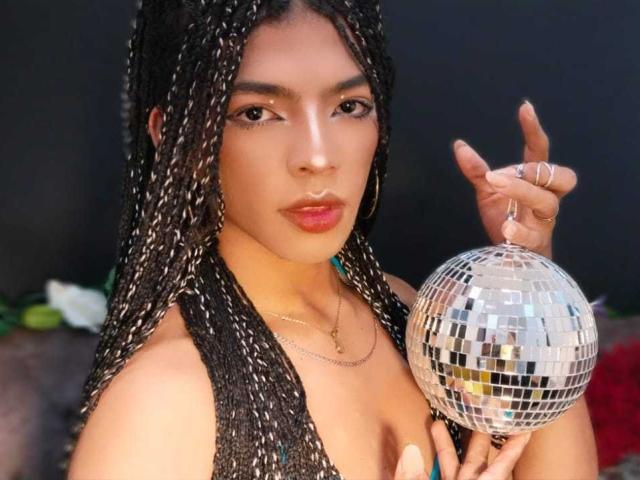 BriyitTs with braided hair holding a mirror ball, wearing a black sleeveless top, has a gold necklace, and a ring, with a serious, slightly pouty expression.