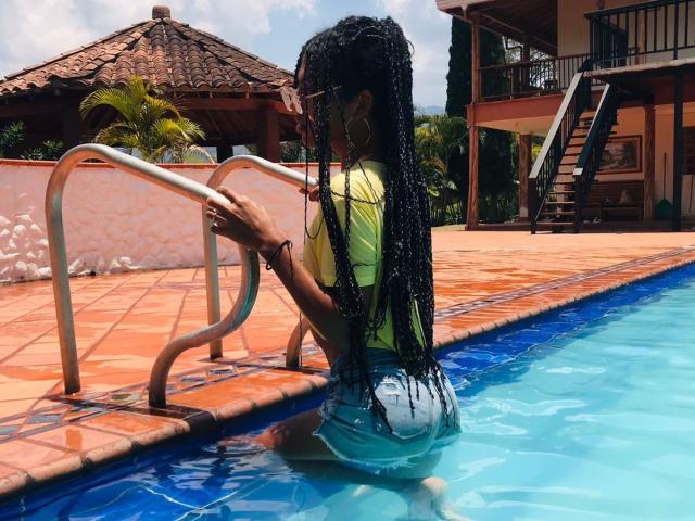 BriyitTs with long black braids, yellow shirt, and denim shorts, kneeling in a pool, gripping metal stairs, blue water, and orange tiled pool deck, wearing hoop earrings.