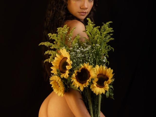 AndromedaZareth's bare ass holding a bouquet of sunflowers and greenery, with curly dark hair, and a golden eye makeup, against a black background.