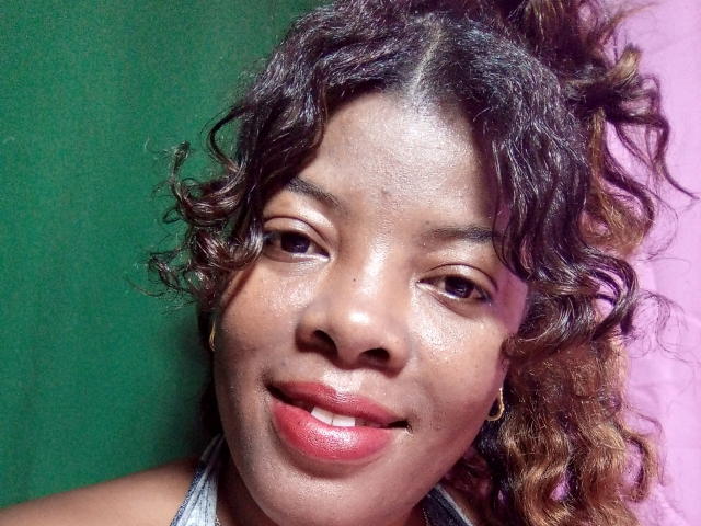 BrownieBabe with curly brown hair, dark brown skin, and red lips, wearing a gray halter top, smiling gently, gold hoop earrings, green and pink background.