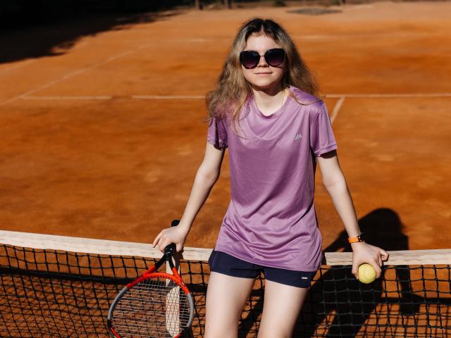 TinaRedly, avec de longs cheveux blonds, portant une chemise violette, un short noir et des lunettes de soleil, tenant une raquette de tennis et une balle, appuyée sur un filet sur un court en terre battue..
