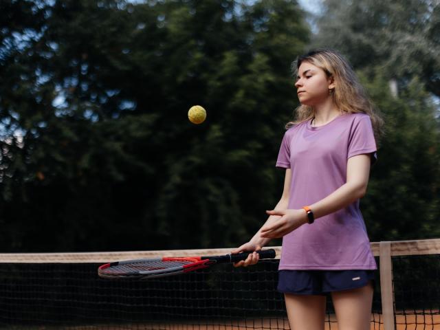 TinaRedly with long brown hair, purple shirt, and dark shorts, holding a tennis racket, eyes on a yellow tennis ball mid-air, standing near a net on an outdoor court.