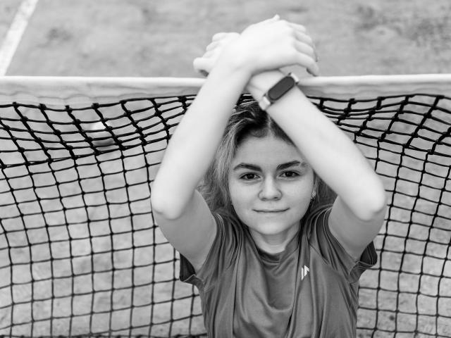 TinaRedly, with arms over net, wearing sports shirt, has a watch on her wrist, and a subtle smile on her face, in black-and-white photo.