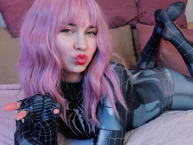 RonyTight with pink hair, pouting lips, wearing black Spider-Man suit, lying on bed, legs up, hand in front, fingers spread, black web pattern, pink bedsheets, cute.