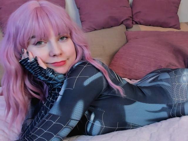 RonyTight with pink hair, lying on bed in black Spider-Man suit, smiling, hand on cheek, showing cute face and red lips, ass slightly raised, looking sexy.