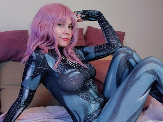 RonyTight with pink hair, wearing a blue Spider-Man suit, sitting on a bed with purple pillows, hand on head, showing her tits and ass, looking at the camera.