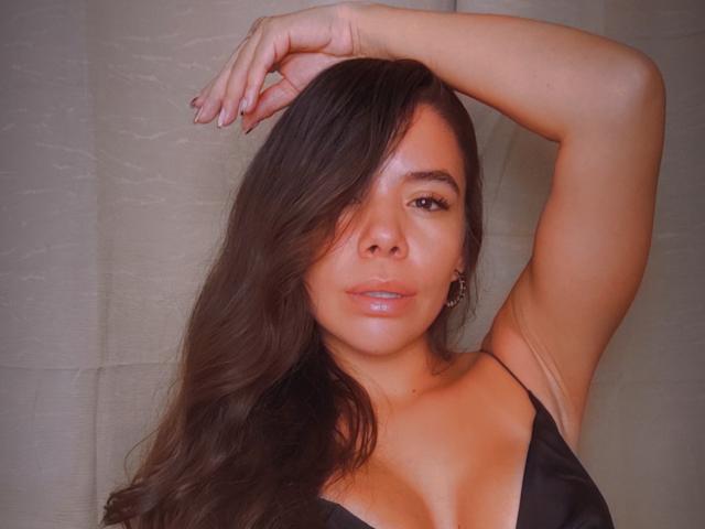 KatiaParker with long brown hair, arm raised, wearing a black dress, showing her tits, glossy pink lips, brown eyes, and subtle makeup, against a plain background.