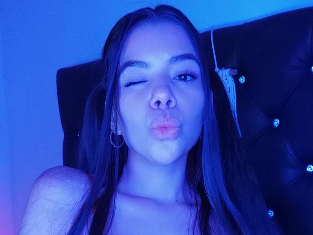 KatiaParker with long dark hair, winking, puckering lips, blue-tinted face, small hoop earrings, black headboard with button-tufted design, wearing a tank top, light skin, slight facial hair.
