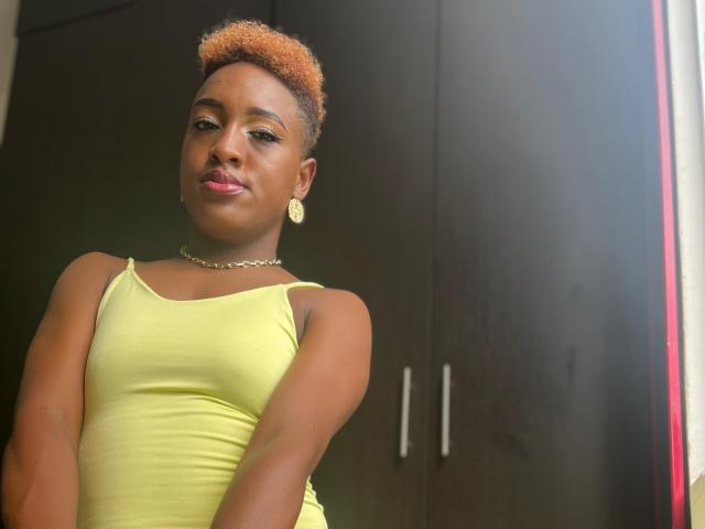 LilaJamez with short orange hair, yellow dress, gold earrings, and necklace, smiling slightly, standing in front of dark wooden cabinets. Her brown skin is smooth, and she's cute.