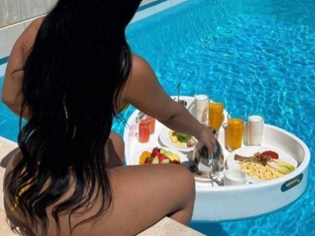 KatyBomb with long black hair, tan skin, and a curvy ass, sits in a pool, eating from a white inflatable tray with drinks, food, and a silver spoon.
