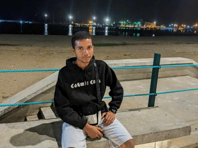 SweetDemonX, short-haired dude in black hoodie and white shorts, sitting on concrete, hands on crotch, rope fence behind, night, city lights reflecting in water, looking at camera.