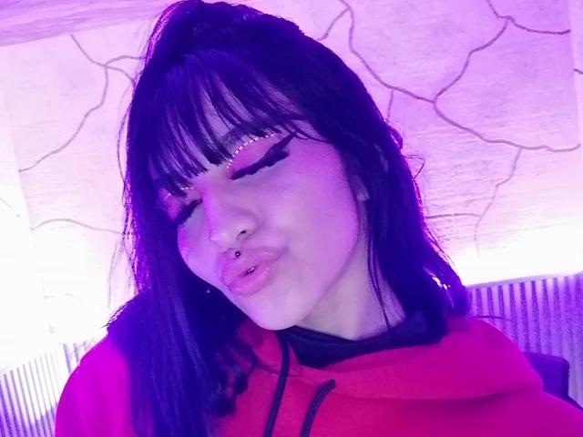 LindaWatsong with black hair, pink eyeliner, and a nose ring, puckering her lips, wearing a red hoodie, illuminated by purple light.
