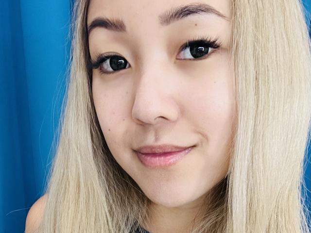 RenyLime's face, blonde hair, dark eyebrows, long eyelashes, slight smile, fair skin, blue eyes, small nose, subtle pink lips, looking at camera, simple background, casual selfie.